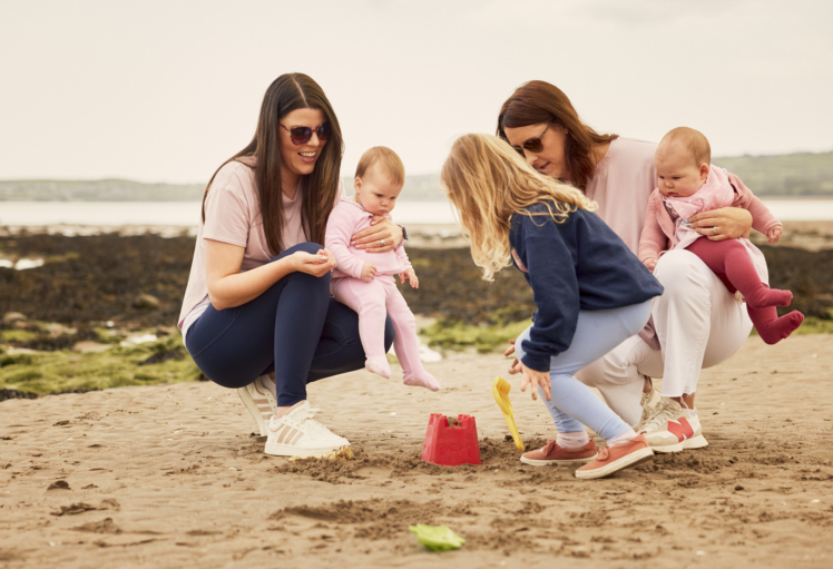 Two mothers on a beach holding toddlers. Mothers are wearing Feed Me Mother Apparel.